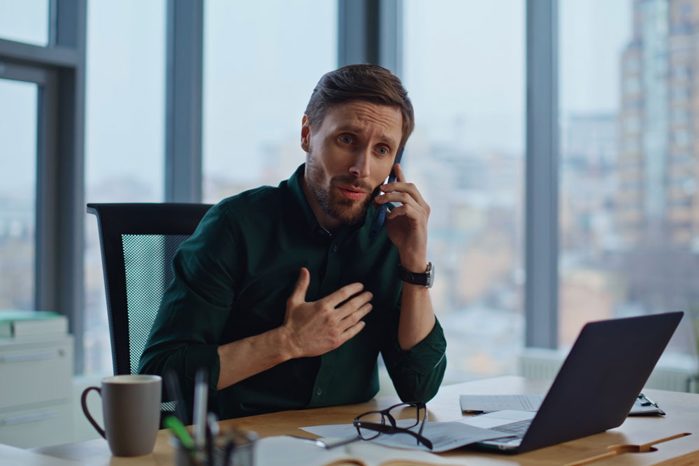A man talking on the phone while sitting at a desk with a laptop in front of him.